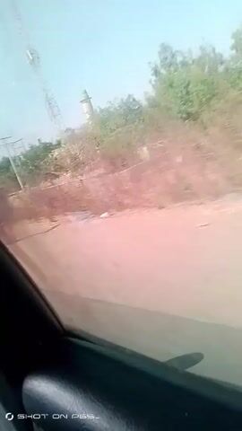 The video is shot from a moving vehicle, possibly a car, traveling along a dirt road. The scenery includes a mosque with a green dome and a tall communications tower. There are trees and a low wall running along the side of the road. Initially, a person wearing red clothing is seen running alongside the road, but they soon disappear from view. The camera then focuses on the passing landscape, including the mosque, tower, and surrounding vegetation. The sky is clear and blue.