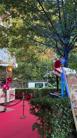 A camera pans right across a festive scene, beginning with a red carpet and velvet ropes leading to a carousel. The carousel is decorated with ornate gold trim, pink flowers, and what appears to be images of roses and poppies. Lights on the carousel are illuminated. Beyond the carousel, a Ferris wheel is visible in the background, partially obscured by trees. A white van is parked further in the background. The scene transitions to a close-up of a sign with floral decorations, featuring Dutch text and the words "ONTDEK DUURBUY." The camera continues to pan right, showing more of the carousel and the surrounding greenery, with blue and purple lights illuminating the trees. The video captures a vibrant, celebratory atmosphere with amusement park elements.