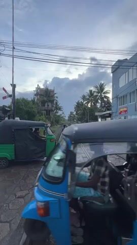This media file captures a street scene in Kodagoda, Sri Lanka, likely during the late afternoon or early evening, given the overcast sky and diffused lighting. The primary subjects are two auto-rickshaws, a mode of transport common in South Asia, and several people. One auto-rickshaw, colored blue, is in the foreground, partially obscuring a second green auto-rickshaw. Both appear to be stopped near a railway crossing.

The setting is a road that runs parallel to a set of train tracks. The road surface is paved with hexagonal stones. In the background, palm trees are visible, indicative of a tropical or subtropical climate. Buildings line the right side of the road, one of which is a light blue, multi-story structure with a sign that appears to read "SHANTHA VILLA" and possibly other text in a local script. The sky is cloudy, suggesting potentially rainy or recently rained-upon conditions, as the road appears wet.

The activity depicted is the movement of people across the train tracks, possibly waiting for or having just passed a train. One person is seen walking along the tracks, and another is approaching them. The auto-rickshaws are positioned near the crossing, suggesting they are either waiting for the barrier to lift or have just passed through. The overall mood is one of everyday life in a busy, yet relaxed, locale. The presence of the railway crossing, overhead electrical wires, and the auto-rickshaws contribute to the urban or semi-urban atmosphere of the location.