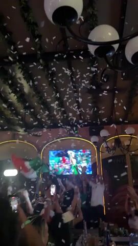 The video shows a ceiling with green vines hanging from it. Two white spherical lights hang from a black chandelier. Below, a crowd of people are gathered, many of them holding up their phones and waving flags. Confetti is falling from above, and a large screen in the background displays a colorful image of a soccer stadium with players. The atmosphere appears celebratory, with people cheering and holding up their devices to capture the moment.