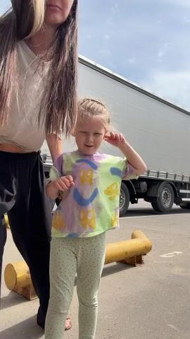 A woman and a small girl are dancing in front of a parked semi-trailer truck. The girl is in the foreground and is wearing a tye-dye shirt and spotted leggings. Her blond hair is braided. The woman is in the background, wearing a beige shirt and black pants. She has long brown hair with blonde highlights. They are standing on an asphalt surface near yellow bollards. The sky is visible above the truck.