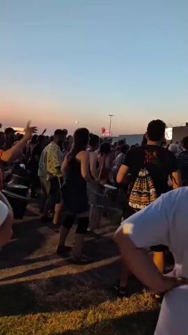 The video captures a lively outdoor event at dusk, possibly a festival or concert, in Alboraia/Alboraya, Spain. A crowd of people are visible, some dancing and others observing. In the foreground, two individuals in white shirts face away from the camera, with one resting their hands on their hips. A group in the mid-ground appears to be engaged in drumming or some form of rhythmic activity, with several individuals holding drumsticks and facing large drums.

The sky is a clear, pale blue transitioning to hues of orange and pink near the horizon, indicating sunset. The overall atmosphere seems energetic and festive, with people enjoying themselves.

In the background, several structures are visible. One building clearly displays the KFC logo in large lettering, suggesting a commercial area near the event. Another building, possibly part of the same complex, has illuminated signs. A tall lamppost is also present. The presence of palm trees in the upper right corner further supports the Spanish coastal location. The lighting suggests it is early evening.