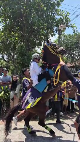 A group of young men, some in matching outfits with purple and yellow accents, are gathered around a horse. One young man, wearing a white shirt and head covering, is seated on the horse. The horse is dark brown, adorned with colorful decorations on its head and body. Several people are holding onto the horse, seemingly to control or support it. The scene appears to be outdoors, possibly a street, with trees and buildings visible in the background under a bright, cloudy sky. Some spectators are also present. The horse is in motion, with its front legs raised.