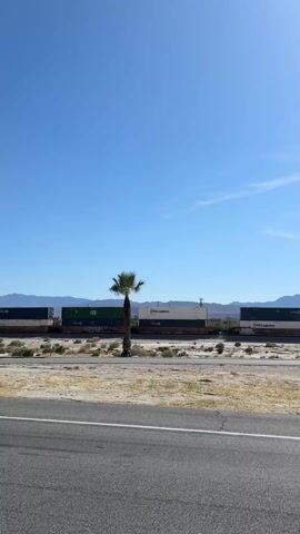 The video shows a freight train with shipping containers moving along a railway track on a sunny day. The sky is clear and blue with a few wisps of clouds. The track runs parallel to a road with a white line marking the edge. A lone palm tree stands between the road and the railway, with desert vegetation in the surrounding area. Mountains can be seen in the distance. The train consists of various containers, some marked with "CTO Logistics" and other company names. The camera is stationary, capturing the scene at eye-level.