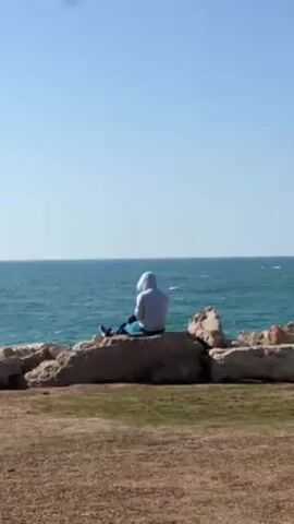 A person wearing a light gray hooded sweatshirt and blue pants sits on a large rock by the sea. The person faces the sea, which has a bright blue color. The sky is cloudless. The rock is part of a formation separating the sea from an area of dry grass. The scene appears to be taken during the day in a place with a clear horizon line.