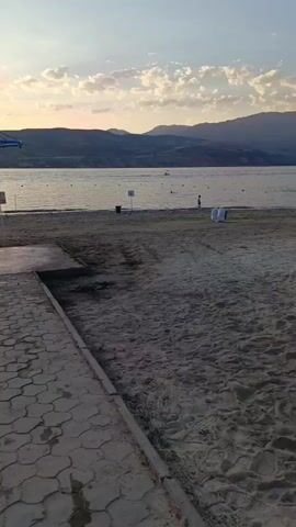 The video shows a wide sandy beach on a body of water. The water is calm and reflects the sky. In the distance, there are hills and mountains. There are a few people on the beach and in the water, some of whom appear to be swimming or playing. There are also some boats visible on the water. The weather appears to be warm and sunny, with some clouds in the sky. The scene is peaceful and relaxing.