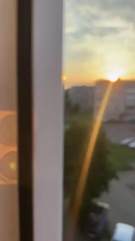 The video captures a sunset view from a window. The sun is visible as a bright orb peeking over a building, casting a golden ray across the scene. In the foreground, a white window frame partially obstructs the view, with a circular lens flare visible on it. The middle ground features a street lined with parked cars and a lush green tree. Beyond the street, apartment buildings dominate the skyline, their windows reflecting the warm hues of the setting sun. The sky is a canvas of soft clouds, tinged with orange and yellow as the sun descends. The overall atmosphere is peaceful and serene.