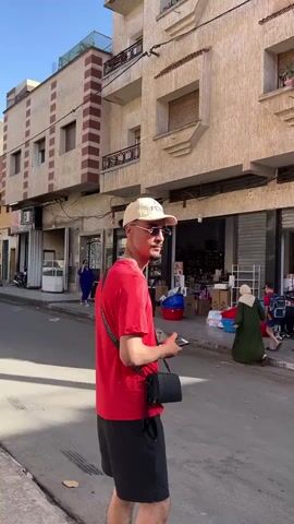 A young man wearing a red shirt, black shorts, a cream-colored baseball cap, and sunglasses is looking at his phone on a street in a Middle Eastern town. He has a black crossbody bag. There are buildings on either side of the street, and people walking in the background. One building appears to be a store, filled with goods. The street is paved and sunny.