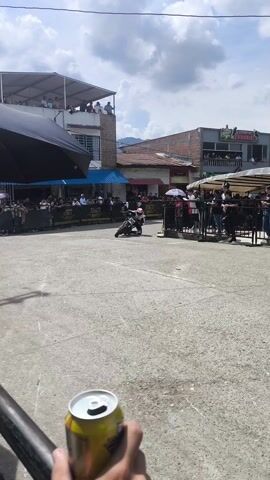 The video shows two motorcyclists performing stunts in a town square. A crowd of people is watching the performance from behind barricades. The motorcyclists are wearing helmets and protective gear. They are riding in circles and leaning low to the ground. A hand holding a yellow and silver beverage can is visible in the lower left corner of the frame. Buildings surround the square and people are watching from rooftops and balconies. The sky is partly cloudy.