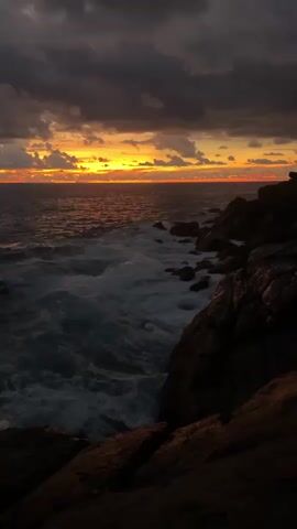 The video depicts a powerful sunset scene over a rocky coastline, likely in Rumassala, Sri Lanka. The sun is setting on the horizon, casting a vibrant orange and yellow glow across the sky, contrasting sharply with the dark, heavy clouds overhead. The ocean is turbulent, with large waves crashing forcefully against the dark, rugged rocks that form the shore in the foreground and mid-ground. White foam from the breaking waves adds dynamism to the deep blue-green water. The rocks appear wet and dark, almost silhouetted against the bright horizon. The overall mood is dramatic and highlights the raw power of nature at dusk. No people or visible text are present in the video.