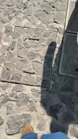 A person walks on a cobblestone road, wearing blue jeans and brown shoes. They are carrying a purse. The road is made of grey, irregular stones, with shadows from the sun and person visible. There are metal sewer covers near the edge of the road.