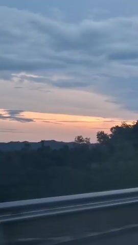 The video shows a view from a moving vehicle looking out at a rural landscape during what appears to be sunset. In the foreground, a grey metal guardrail is visible, suggesting the viewpoint is from a road. Beyond the guardrail, there is a stretch of dark green foliage, likely trees and bushes, that line a gently sloping hill. In the distance, faint outlines of mountains can be seen under a sky filled with soft, grey clouds tinged with hues of orange and pink from the setting sun. The lighting suggests dusk or early evening.