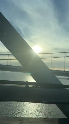 The video shows a view from a moving vehicle crossing a large suspension bridge. The sun is bright and low in the sky, creating a strong glare and lens flare, partially obscuring the view. The bridge structure, including cables and support beams, is visible. The water below the bridge reflects the sunlight. The sky is partly cloudy with wispy clouds. Distant lights and a few other vehicles can be seen on the bridge. The video is filmed from inside the vehicle, with the dashboard or interior elements visible in the foreground.