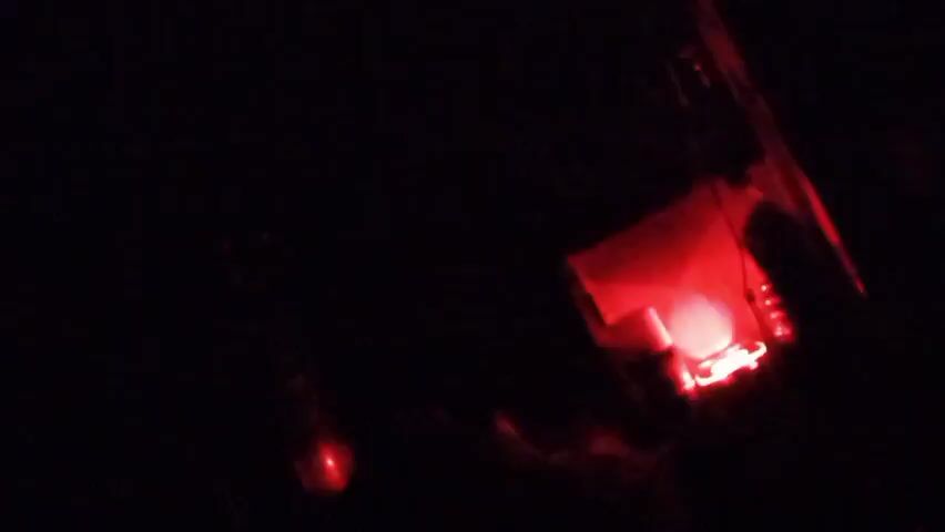 The video shows a small, dark, possibly cubic object emitting lights in various colors – red, blue, yellow, and purple – against a completely black background. The light is sporadic and doesn’t uniformly illuminate the object, making it difficult to discern specific features. The object is in the lower right portion of the frame.