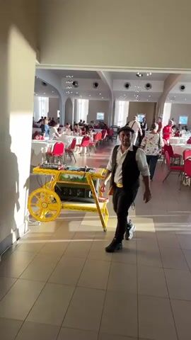 A man wearing a flat cap, a white shirt, a dark vest, and black pants is dancing in front of a yellow cart with decorative wheels. The cart appears to be a DJ booth or music player. The man is in a large, well-lit room with high ceilings. In the background, tables with red tablecloths are set up, and people are seated, eating or socializing. Sunlight streams in from the left, casting shadows on the floor and wall. The man is animated, with his arms outstretched and one leg raised as he dances. He turns and gestures towards the wall, then back towards the cart, seemingly engaging with the audience. The overall atmosphere appears to be a celebratory event or gathering.