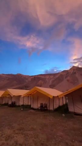 The video shows a panning view of a campsite at dusk. Several white tents with yellow roofs are arranged in a row. Behind the tents, a mountainous landscape is visible. The sky is filled with dramatic clouds painted in shades of pink, orange, and blue, suggesting either a sunrise or sunset. The ground in front of the tents appears to be a dirt or gravel surface. The camera movement is smooth, indicating a deliberate sweep across the scene. There are no people visible in the video. The overall atmosphere is serene and picturesque.