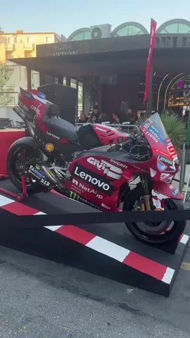 A red and black Ducati motorcycle with "Lenovo" and "Givi" branding is displayed on a ramp. The motorcycle is a racing bike. People are visible in the background under a structure with "Tequila Patron" signage. The environment appears to be an outdoor setting, possibly a bar or restaurant with a patio.