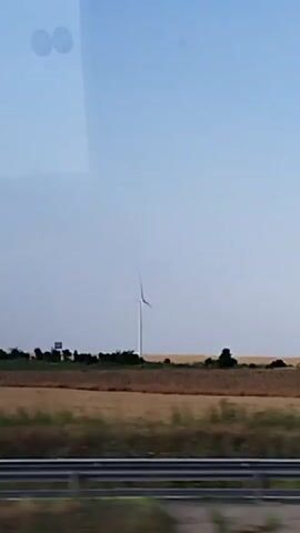 A wide shot shows a rural landscape under a clear blue sky. In the middle distance, a lone white wind turbine stands tall, its blades stationary. A flat field of golden-brown crops stretches across the frame, bordered by a line of dark green trees in the background. In the foreground, a blurred section of a grey railing runs horizontally across the bottom of the image, suggesting the viewer is looking from a moving vehicle. The overall impression is one of a vast, open countryside.