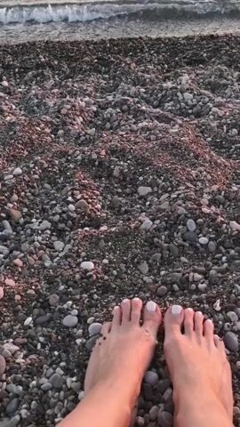 The video shows a pair of human feet on a pebble beach. The feet are positioned facing the camera and are painted with white nail polish. The pebbles are dark gray, brown, and black, with some reddish-brown ones mixed in. In the background, the ocean is visible with gentle waves breaking on the shore. The lighting suggests it is either sunrise or sunset.