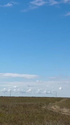 The video shows a wide shot of a flat, grassy field under a bright blue sky with scattered white clouds. Two thin, dark poles are visible in the distance, standing vertically on the horizon. A dirt path winds through the field, leading towards the horizon and between the two poles. The grass in the field is dry and brown in some areas, with some green patches. The lighting suggests it is daytime. There are no people or animals visible in the video. The camera is stationary, and the scene is peaceful and natural.