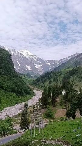 A view of a mountain valley with a river and a road. The mountains are covered in green trees and snow-capped peaks. There are several cars driving on the road. The sky is cloudy.