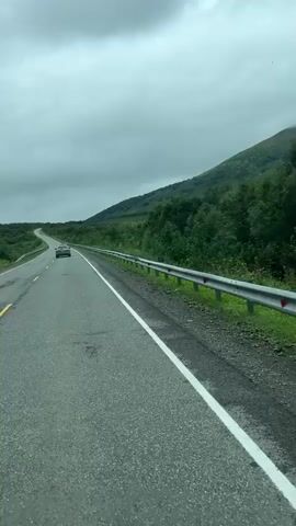 The video captures a view from a vehicle traveling on a paved road. The road curves gently to the left and is bordered by a guardrail on the right. Lush green vegetation covers the rolling hills to the right of the road, with a larger, mist-shrouded mountain visible in the background. A single car is visible in the distance, traveling in the same direction. The sky is overcast with thick, gray clouds, suggesting a cool or possibly rainy day. The overall scene depicts a scenic, natural landscape encountered during a road trip.