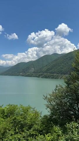 This is a vertical, wide shot of a vast, turquoise lake surrounded by lush green mountains under a bright blue sky dotted with fluffy white clouds. The sun is shining, indicating it is daytime, likely mid-morning or early afternoon, given the clear visibility and vibrant colors. The scene is natural and serene, conveying a sense of peace and beauty.

The foreground is filled with dense green foliage, suggesting a viewpoint from a hillside or elevated area overlooking the lake. The lake itself is the dominant subject, its calm surface reflecting the sky and mountains. The mountains in the background are rolling and covered in trees, with a winding road visible along the edge of the lake in the middle distance, hinting at the presence of human activity in the area. The overall impression is one of a remote and picturesque natural landscape.

The location is identified as Aranisi, Georgia. The image captures a tranquil moment in nature, with no people or specific objects visible, focusing entirely on the expansive scenery. The weather appears to be pleasant and fair, conducive to outdoor appreciation.
