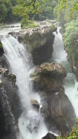 This video shows a powerful waterfall cascading over rocks in a gorge. Water flows from left to right, creating a dramatic scene with lush green trees and foliage surrounding the rocky cliffs. The water is white and foamy as it plunges down, creating mist and spray. The overall impression is one of nature's raw power and beauty.