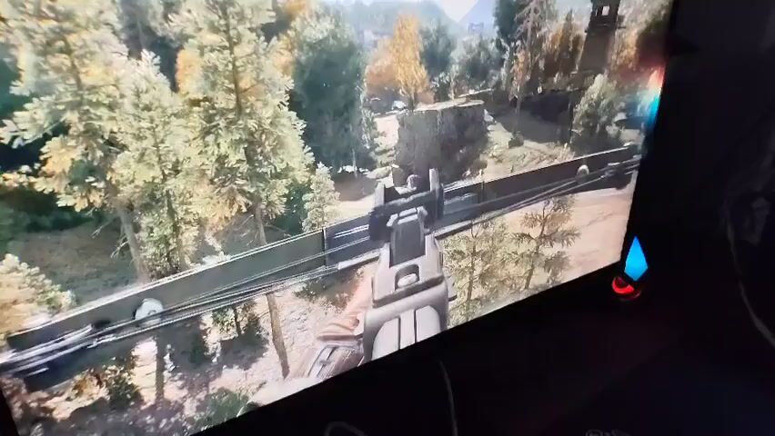 This media file captures a first-person perspective of a video game, likely a first-person shooter or action-adventure, being played on a computer monitor. The game's graphics depict a dense forest scene with tall evergreen trees and some deciduous trees showing autumn foliage in shades of yellow and orange. In the foreground, the player's in-game character is holding a crossbow, which is prominently displayed across the bottom half of the screen. The crossbow appears to be loaded, with arrows nocked and ready.

In the background, amidst the trees, there are remnants of man-made structures, including what looks like a small, weathered tower or lookout post on a hill. There are also hints of rocky terrain and possibly distant mountains. The lighting suggests it is daytime, with sunlight filtering through the trees, creating a natural and somewhat rustic atmosphere. The game's visual style appears realistic, aiming for an immersive experience.

The monitor itself is visible at an angle, with a dark bezel and some illuminated elements on its right side, suggesting it's an LED-lit monitor with dynamic lighting features. The scene from the game evokes a sense of wilderness and perhaps impending action, given the weapon being held. No people are visible in the frame, except for the character's gloved hand holding the crossbow. The weather appears clear and pleasant, conducive to outdoor activity. There is no visible text within the game's scene or on the monitor frame.