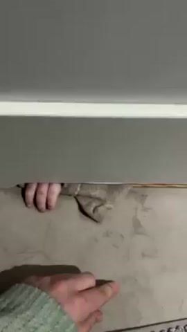 A short video shows a hand reaching under a gray surface, possibly a cabinet or shelf, taking a gray rag away. The hand then goes back to reaching under the gray surface, then withdraws again and picks up the rag.