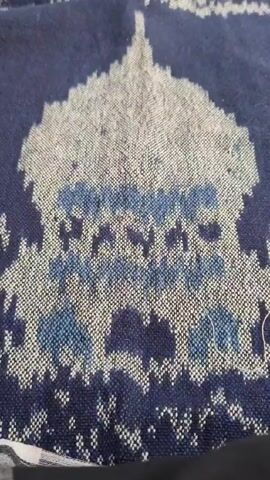Here is a description of the video:

The video shows a man praying on a prayer rug with a mosque design in what appears to be an outdoor or semi-outdoor setting. The camera starts focused on the prayer rug itself, showcasing its intricate pattern and colors. The rug is primarily blue with a mosque design woven in lighter shades, including white and blue, and is sitting on a wooden surface. The man, wearing a white shirt, green pants, and a white prayer cap, kneels on the rug, facing away from the camera. The surrounding environment includes plants and a glimpse of a building in the background.