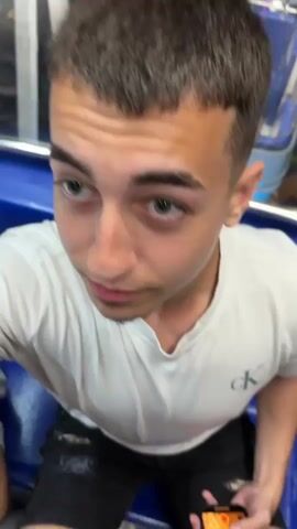 A teenager in a white shirt is sitting in a Ferris wheel cabin at night, looking upwards and slightly to the left. The cabin has a blue interior with metal railing. The wheel is illuminated with multicolor lights on the frame, rotating past other cabins. The background shows an urban landscape at night with buildings and streetlights. Music is audible.