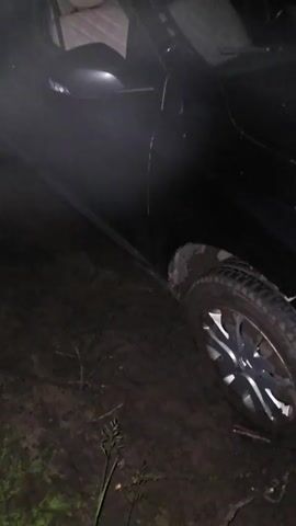 A black car is stuck in mud at night. The car's interior is visible, showing beige seats. A utility pole stands to the right of the car, with grass and weeds growing around its base. The scene is dark, with limited visibility.