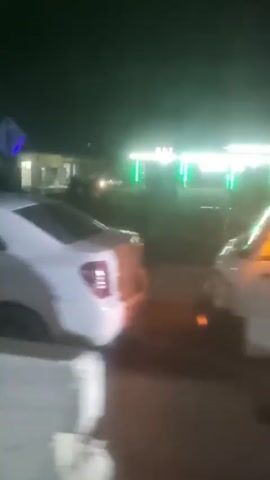 At night, a person in a dark hoodie and blue jeans walks past a white car. The car is parked near a white van, and there are lights from a building behind them. The camera moves past the car and shows more of the street. Another car, yellow, is seen parked on the side of the road.
