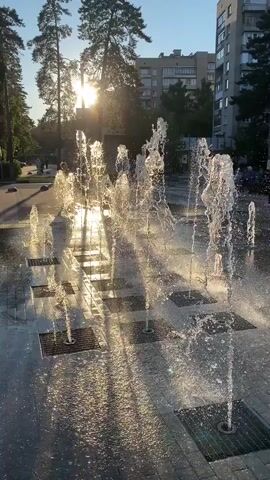 The video shows a dynamic fountain installation with multiple water jets spraying upwards from a paved surface. The sun shines brightly, casting a warm glow and creating lens flares. Several people are visible in the background, walking and observing the fountain. Some children appear to be interacting with the water. The environment suggests an urban park or public square, with trees and apartment buildings in the background. The overall atmosphere is lively and recreational.