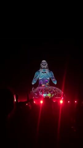 A holographic projection of a humanoid figure is displayed against a black background. The figure appears to be a stylized representation of a mythical creature, possibly a mermaid or a water spirit, with elements of aquatic life such as fish and coral projected onto its body. The figure is seated on a transparent, dome-like structure that is illuminated with vibrant red and pink lights, creating a pulsating effect. Beneath the dome, a crowd of people is visible, their silhouettes outlined by the red lighting. Many individuals are holding up their phones, presumably to record the spectacle. The overall atmosphere suggests a nighttime event or performance, likely a concert or festival, characterized by dramatic lighting and visual effects.