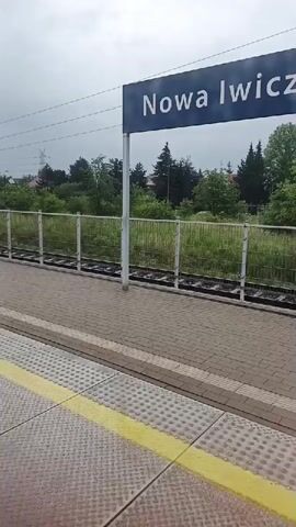 The video shows a train station platform with a sign that reads "Nowa Lwczna". The platform is paved with bricks and has a yellow tactile warning strip along the edge. A metal fence separates the platform from the train tracks, beyond which there is tall grass and trees. The sky is overcast.