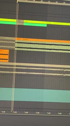A close-up shot shows a computer screen displaying a digital audio workstation (DAW) software interface. Several audio tracks, represented by colored blocks (green, orange, gray, light gray, and teal), are visible in the arrangement view of the DAW. A vertical playhead line moves across the screen, indicating the current playback position. The timeline at the bottom shows time markings from 1:16 to 1:30.  The software interface appears complex with multiple waveforms and audio clips, suggesting a music production or audio editing task.