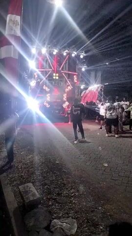 A group of people stand in front of a large sound system with flashing lights. The sound system is set up on a stage or platform with multiple speakers and lights. Some people are holding flags, and others are standing and observing the scene. The ground appears to be paved with cobblestones. The lighting suggests it is nighttime or a dimly lit area.