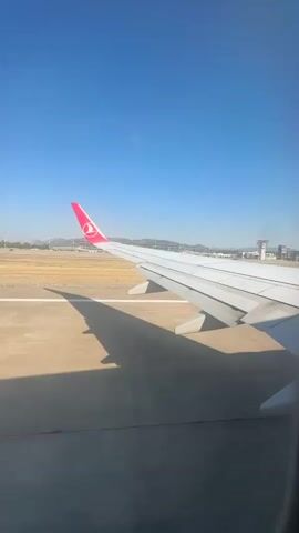This is a first-person perspective video taken from an airplane window during daytime. The scene captures the airplane's wing and its shadow on the tarmac. The tail of the airplane, with a red logo, is visible. The runway and airport infrastructure, including buildings and control towers, are in the background. The sky is clear and blue. The plane appears to be moving on the ground, likely taxiing or accelerating for takeoff.