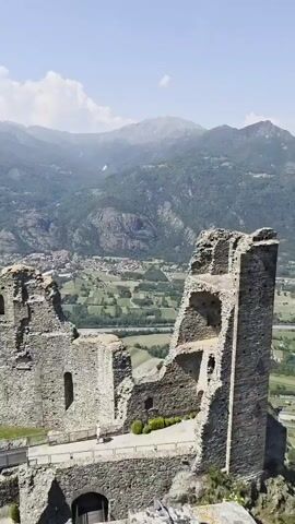 The video shows a partially ruined stone castle with mountains in the background on a bright, sunny day with some clouds.  There is grass within the walls and a walking bridge, with two people walking on the bridge. A town can be seen in the valley below. The camera pans to show more of the town and valley below, as well as the surrounding mountains.