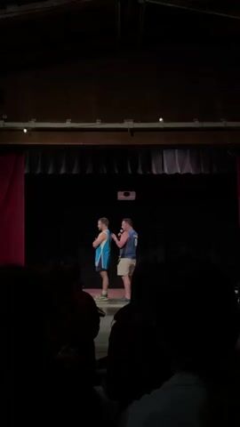 Two men stand on a dark stage. The man on the left wears a blue and white basketball jersey and black shorts. The man on the right wears a dark blue tank top with the number 0 on it and khaki shorts, and holds a microphone. He appears to be speaking to the man on the left, who looks away. The stage has a dark curtain on the left and a projector mounted on the ceiling. The foreground is obscured by the silhouettes of audience members.