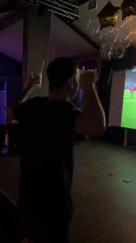 A person in a dark t-shirt stands with their back to the camera, facing a large projection screen in a dimly lit room. The screen displays a soccer match, showing two men in soccer jerseys embracing on a field. One man is wearing a blue and black jersey with the name "MESSI" and the number "10" on the back. Above the screen, the word "BRAVA" is illuminated in large, orange, block letters. Several gold star-shaped balloons and clear, round balloons are clustered near the ceiling. The floor is made of dark wood.