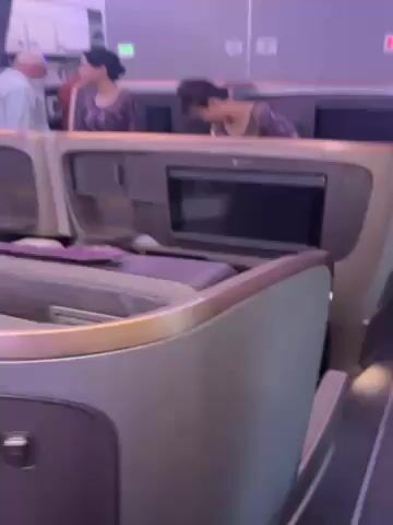 The video shows the interior of an airplane, specifically the first-class section. The seating area is seen with dividers providing privacy. Each seat has a built-in entertainment screen.  Two flight attendants in patterned uniforms are visible; one seems to be preparing a seat, while the other stands further away. A man in a white shirt is partially visible in the background. A person wearing a black jacket, white shirt, black pants, and white sneakers is walking down the aisle. The interior lighting has a purplish hue.