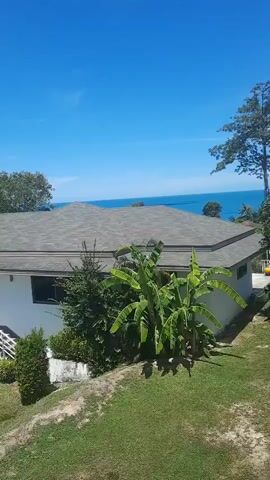 The video shows a panning view of a tropical landscape with a clear blue sky and white clouds. In the foreground, there is a grassy slope with some manicured bushes. A white building with a gray roof is visible, featuring an external staircase leading to a door. Banana trees and other lush green vegetation surround the building. In the background, the ocean can be seen, and further in the distance, there are rolling hills covered in dense forest. The camera pans from left to right, revealing more of the wooded hills and another white building with a circular window feature, set amidst the greenery. The scene appears to be a scenic view from a higher vantage point, possibly a balcony or a hill. There are no people or animals visible in the video.