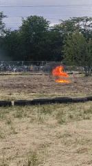 Silver car burns in grassy area as crowd watches
