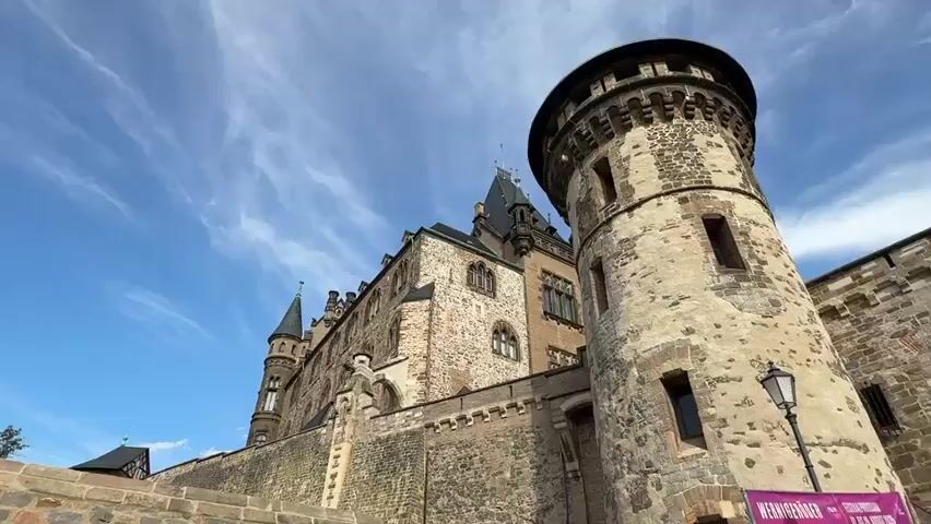 The video shows an upward view of a castle made of stone and brick. The castle has several towers and turrets, some of which are round and others are square. The architecture appears to be medieval or Gothic, with pointed arches and intricate detailing on the windows and roofs. The sky is blue with some wispy clouds. The video pans across the facade of the castle, highlighting its imposing structure and weathered stone. There are no people or other objects visible in the frame.