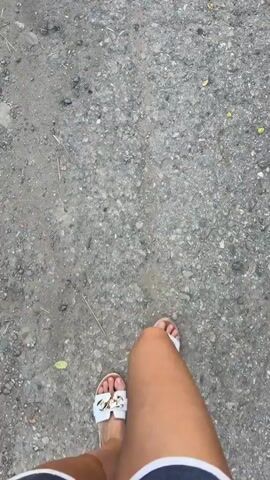 A POV video shows a person walking down a dirt road. The person's feet are visible wearing white sandals. The road is rough and appears to be in a rural area with trees and vegetation on either side. In the distance, there are a few cars and houses visible. The sky is cloudy.
