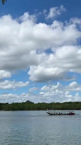 A long, narrow boat with a red and white canopy is moving across a body of water. Several people are seated inside the boat, which appears to be a passenger vessel. The water is calm, and the sky is a bright blue with scattered white clouds. In the background, a dense line of green trees forms a shoreline, with a small hill visible behind the trees on the left. The overall scene depicts a tranquil day on the water, likely in a tropical or coastal environment.