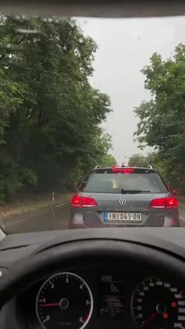 The video is a first-person perspective from inside a car, looking through the windshield which is speckled with raindrops. The car is in traffic on a paved road lined with dense green trees and foliage. The steering wheel and part of the dashboard are visible in the foreground. Ahead, a gray Volkswagen Passat is stopped in traffic, its brake lights illuminated. The license plate is partially visible. To the right, another vehicle, possibly a van, is in the adjacent lane. The weather appears overcast and rainy, as indicated by the raindrops on the windshield and the general lighting conditions.